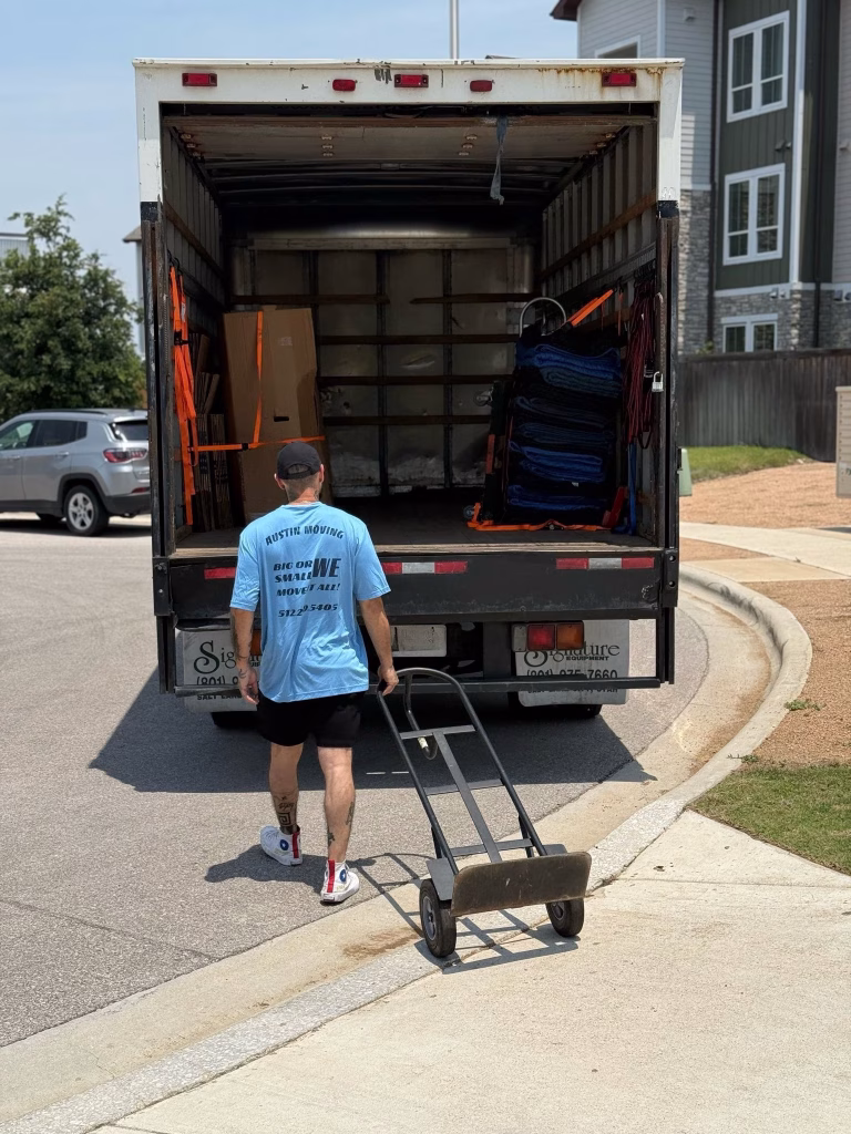 A professional mover pushing an empty hand truck toward an open moving truck loaded with moving blankets.